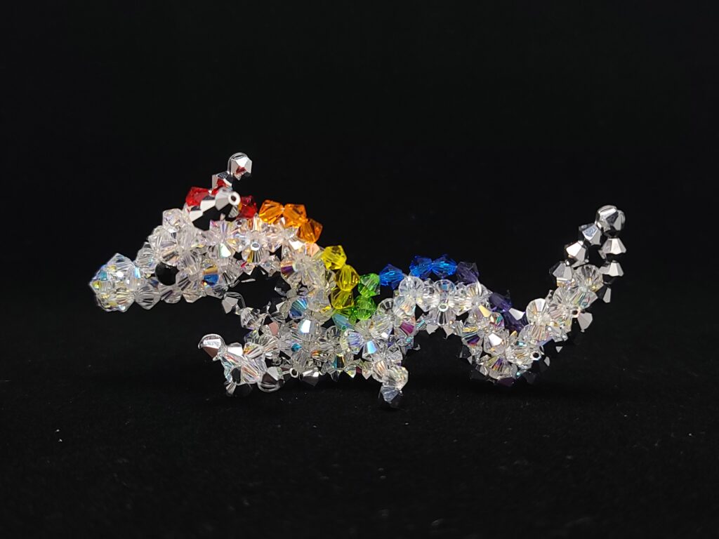 Een klein beeldje van een draak gemaakt met kristallen kralen tegen een zwarte achtergrond. De draak is een Aziatische draak. Het lichaam is wit, de ogen zwart, en de draak heeft manen in felle regenboogkleuren. De hoorns, klauwen en de buik van de draak zijn zilver. A small beaded dragon figurine against a black background. The dragon is an Asian dragon, with a white body, black eyes, manes consisting of bright rainbow colours and silver horns/claws/belly.