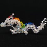 Een klein beeldje van een draak gemaakt met kristallen kralen tegen een zwarte achtergrond. De draak is een Aziatische draak. Het lichaam is wit, de ogen zwart, en de draak heeft manen in felle regenboogkleuren. De hoorns, klauwen en de buik van de draak zijn zilver. A small beaded dragon figurine against a black background. The dragon is an Asian dragon, with a white body, black eyes, manes consisting of bright rainbow colours and silver horns/claws/belly.