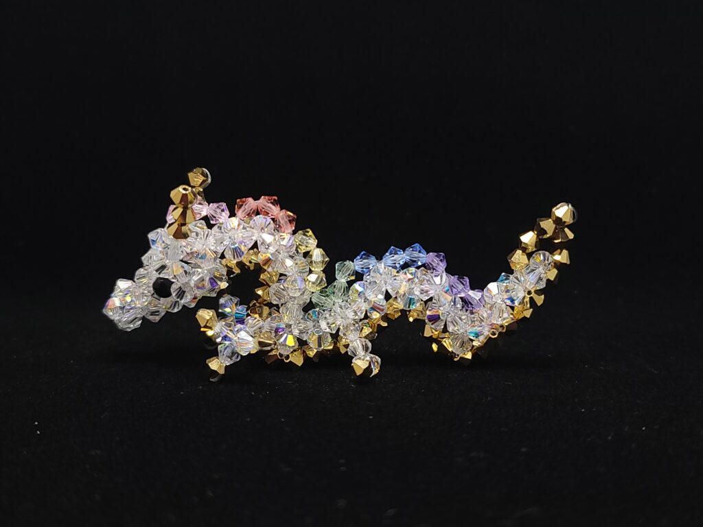 Een klein beeldje van een draak gemaakt met kristallen kralen tegen een zwarte achtergrond. De draak is een Aziatische draak. Het lichaam is wit, de ogen zwart, en de draak heeft manen in pastel regenboogkleuren. De hoorns, klauwen en de buik van de draak zijn goud. A small beaded dragon figurine against a black background. The dragon is an Asian dragon, with a white body, black eyes, manes consisting of soft pastel rainbow colours and gold horns/claws/belly.