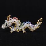 Een klein beeldje van een draak gemaakt met kristallen kralen tegen een zwarte achtergrond. De draak is een Aziatische draak. Het lichaam is wit, de ogen zwart, en de draak heeft manen in pastel regenboogkleuren. De hoorns, klauwen en de buik van de draak zijn goud. A small beaded dragon figurine against a black background. The dragon is an Asian dragon, with a white body, black eyes, manes consisting of soft pastel rainbow colours and gold horns/claws/belly.