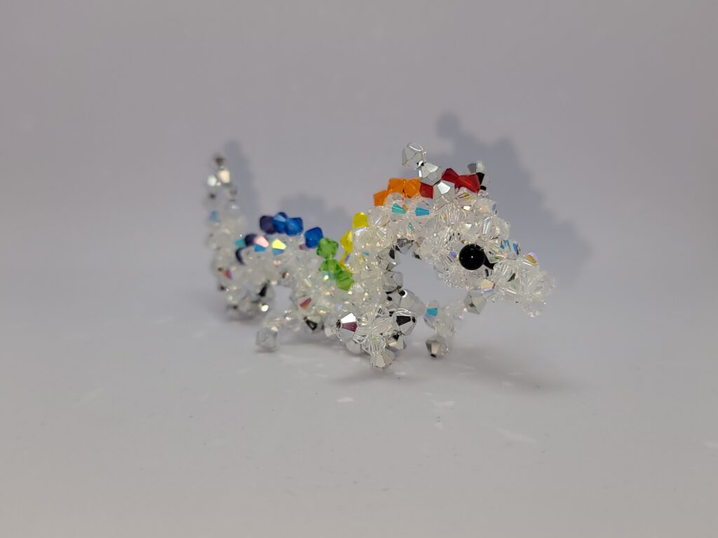Een klein beeldje van een draak gemaakt met kristallen kralen tegen een zwarte achtergrond. De draak is een Aziatische draak. Het lichaam is wit, de ogen zwart, en de draak heeft manen in felle regenboogkleuren. De hoorns, klauwen en de buik van de draak zijn zilver. A small beaded dragon figurine against a black background. The dragon is an Asian dragon, with a white body, black eyes, manes consisting of bright rainbow colours and silver horns/claws/belly.