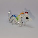 Een klein beeldje van een draak gemaakt met kristallen kralen tegen een zwarte achtergrond. De draak is een Aziatische draak. Het lichaam is wit, de ogen zwart, en de draak heeft manen in felle regenboogkleuren. De hoorns, klauwen en de buik van de draak zijn zilver. A small beaded dragon figurine against a black background. The dragon is an Asian dragon, with a white body, black eyes, manes consisting of bright rainbow colours and silver horns/claws/belly.